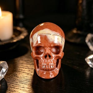 May include: A polished, reddish-brown skull figurine with a cream-coloured patch on top. The skull has detailed features, including eye sockets, a nose cavity, and teeth. The figurine is likely made of stone or a similar material, and is a decorative item.