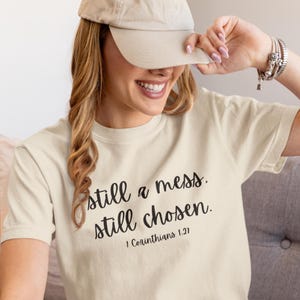 May include: Cream-colored t-shirt with the text "still a mess. still chosen." and the Bible verse 1 Corinthians 1:27 in black script. The person in the image is wearing a beige baseball cap.