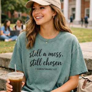 May include: Sage green t-shirt with the text "still a mess, still chosen" and "1 Corinthians 1:27" in black script. The shirt is worn with a beige baseball cap and a gold necklace. A clear plastic cup with a straw is held in the hand.
