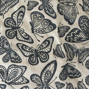 May include: A beige fabric with a repeating pattern of black butterflies. The butterflies have intricate designs and are arranged in a random pattern.