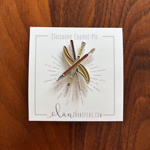 May include: A cloisonné enamel pin featuring crossed paintbrushes and a palette design. The pin has silver, brown, and yellow accents, with the text "Cloisonné Enamel Pin" at the top. The website elantransfers.com is printed at the bottom.