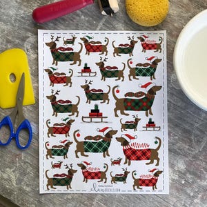 Puede incluir: Una hoja de pegatinas de Navidad con perros salchicha. Las pegatinas presentan perros salchicha marrones con suéteres, sombreros y cornamentas festivas. Algunos perros tiran de trineos o llevan regalos. Las pegatinas están sobre un fondo blanco.