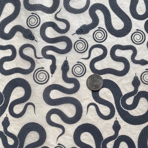 May include: A white fabric with a repeating pattern of black snakes. The snakes are stylized and have a textured appearance. The fabric has a subtle, off-white background.