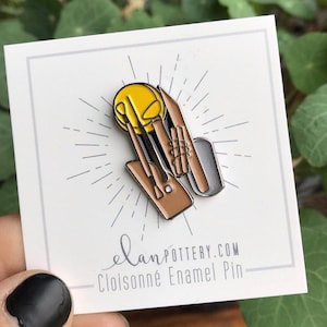 May include: A cloisonné enamel pin featuring a yellow circle and brown pottery tools. The pin has a silver and brown metallic finish. The pin is on a white card with the text "elanPOTTERY.COM Cloisonné Enamel Pin".
