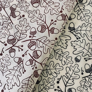 May include: Two pieces of fabric with a repeating pattern of brown and black oak leaves and acorns. The fabric on the top is brown and the fabric on the bottom is black.