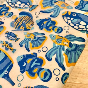 Könnte beinhalten: Nahaufnahme einer gemusterten Oberfläche mit stilisierten Fischillustrationen in Blau- und Gelbtönen. Die Fischdesigns umfassen Streifen, Punkte und wellenförmige Muster, wobei einige Fische lange, fließende Flossen haben. Weiße Blasen sind über das Design verstreut.