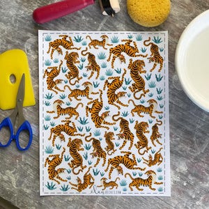 May include: A white sheet with a repeating pattern of orange and black tigers interspersed with blue-green foliage. The sheet is surrounded by craft tools, including a yellow scraper, scissors, and a red roller.