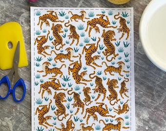 May include: A white sheet with a repeating pattern of orange and black tigers interspersed with blue-green foliage. The sheet is surrounded by craft tools, including a yellow scraper, scissors, and a red roller.