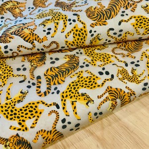 May include: A fabric featuring a repeating pattern of orange and black striped tigers and yellow and black spotted leopards on a light cream background. The wild cats are depicted in various dynamic poses, interspersed with scattered black spots.