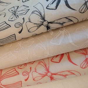 May include: Three rolls of wrapping paper with bow designs. The top roll has black bows, the middle roll has white bows, and the bottom roll has red bows. The paper has a kraft paper appearance.