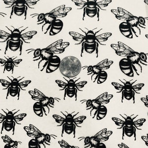 May include: A cream-colored fabric with a repeating pattern of black bees. The bees are depicted in a detailed, stylized illustration, with visible wings and body segments. A quarter is included for scale.