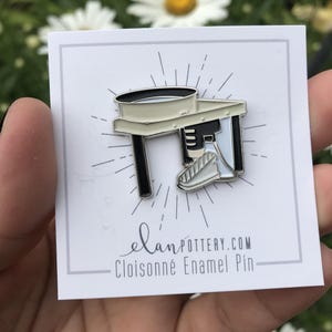 May include: Cloisonné enamel pin featuring a pottery wheel design. The pin has a black and white color scheme with a light beige wheel and table, and black legs. The pin is on a white card with the text "elanpottery.com Cloisonné Enamel Pin".