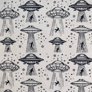 Puede incluir: Patrón repetido con ilustraciones en blanco y negro de ovnis, cada uno emitiendo un rayo de luz que parece abducir una figura humana. El fondo es de color beige claro con formas de estrellas dispersas.