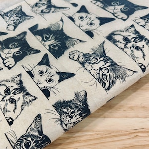 May include: A beige fabric with a repeating pattern of stylized cat faces in dark blue ink. The cats are depicted in various poses, peeking out from behind a border. The design has a hand-drawn, whimsical quality, suitable for crafting or home decor projects.