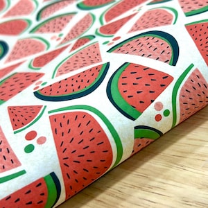 May include: White paper with a repeating pattern of watermelon slices. The watermelons are red with black seeds, green rinds, and dark blue outlines. The paper is slightly curled, revealing the pattern's continuity.
