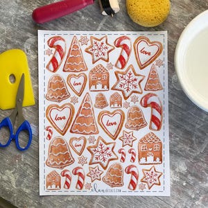 May include: A sheet of watercolor-style gingerbread cookie stickers. The stickers include gingerbread houses, stars, hearts with the word "love", candy canes, bells, and Christmas trees. The stickers are in shades of brown and red.
