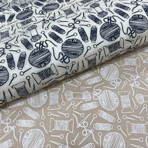 Puede incluir: Tela con un patrón repetido de artículos de costura. El diseño presenta carretes de hilo, ovillos de lana, agujas y tijeras en azul oscuro sobre un fondo crema. Se ve una segunda capa de tela con un diseño similar en blanco sobre un fondo beige.
