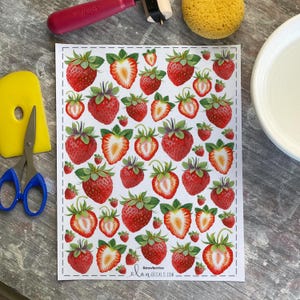 May include: A sheet of strawberry decals with a white background. The decals feature red strawberries with green leaves, some whole and some sliced. The text "Strawberries" and "elanDECALs.com" is printed on the sheet. Craft supplies are in the background.