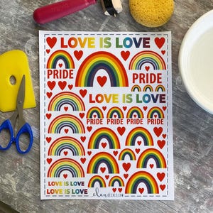 May include: A white sheet with a repeating pattern of rainbow arches, red hearts, and the words "LOVE IS LOVE" and "PRIDE" in a colorful design. The sheet is surrounded by crafting tools.