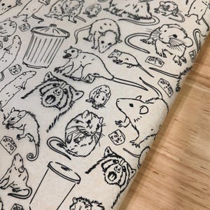 Puede incluir: Tela blanquecina con un patrón repetido de dibujos lineales negros de varios roedores, incluyendo ratas, zarigüeyas y hámsteres. El diseño también incluye botes de basura y cuñas de queso. La tela está doblada, revelando una superficie de madera.
