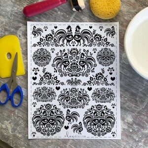 May include: Black and white decorative decal sheet featuring stylized roosters, floral patterns, and hearts. The sheet includes the text "elan decals.com" at the bottom. A yellow tool, scissors, a red roller, and a sponge are visible.