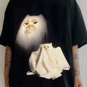 Pu&ograve; includere: T-shirt nero con un disegno di gatto bianco dall'aspetto spettrale. Il gatto ha segni facciali gialli e marroni. Il disegno &egrave; stampato sul davanti. La maglietta ha maniche corte e girocollo.