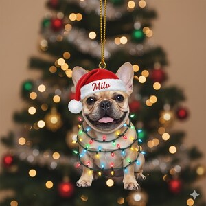 May include: A Christmas ornament featuring a smiling French Bulldog wearing a Santa hat with the name "Milo" and a string of colorful Christmas lights. The ornament hangs in front of a blurred Christmas tree.