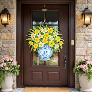 May include: A decorative door wreath featuring a vase filled with yellow and white daffodils. The blue and white vase has the letter "H" on it. The wreath hangs on a brown door with a "JOY" sign.