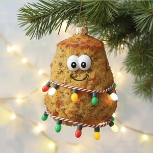 May include: A whimsical Christmas ornament shaped like a baked potato, adorned with a smiling face and a string of colorful lights. The ornament is suspended from a gold-colored cap and a green pine branch.