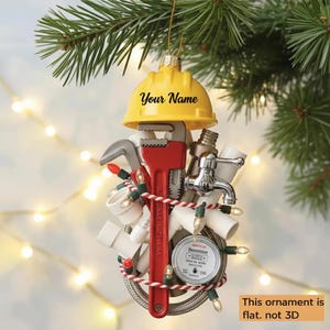 May include: A yellow hard hat Christmas ornament with the text "Your Name". The ornament is decorated with plumbing tools, a red wrench, and a string of lights. The ornament is hanging from a Christmas tree branch.