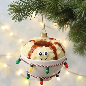 May include: A whimsical Christmas ornament shaped like a bowl of mashed potatoes and gravy, with a smiling face and string lights. The ornament is white with a gold-colored top and a red and white striped trim.