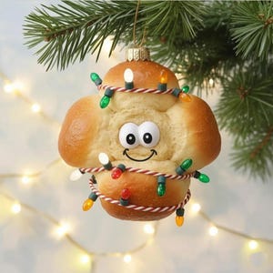 May include: A whimsical Christmas ornament shaped like a smiling baked good, adorned with a string of colorful lights. The ornament is suspended from a gold-colored cap and a green pine branch, creating a festive holiday decoration.