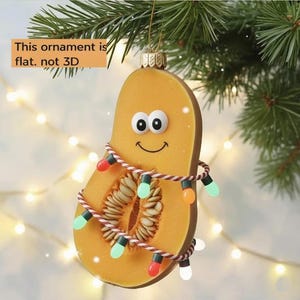 May include: A flat, pumpkin-shaped Christmas ornament with a smiling face and googly eyes. The ornament is wrapped with a string of colorful lights. The text on the image says: "This ornament is flat. not 3D."