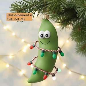 May include: A green pea pod Christmas ornament with a smiling face and wrapped in a string of colorful lights. The ornament is hanging from a gold-colored cap and a pine branch. Text on the image says: "This ornament is flat. not 3D."