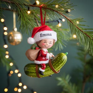 May include: A Christmas ornament featuring a smiling figure in a Santa hat with the name "Emma" riding a pickle. The figure wears a red suit and carries a sack of gifts. The ornament hangs from a red ribbon on a Christmas tree.
