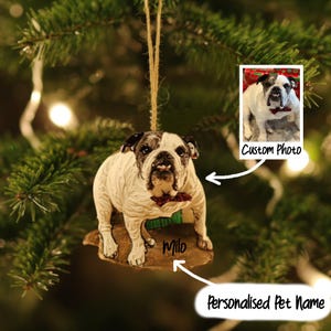 May include: A white and brown bulldog ornament with a red bow tie and the name "Milo". The ornament is hanging from a Christmas tree. A small photo of the dog is in the upper right corner. Text reads "Custom Photo" and "Personalised Pet Name".