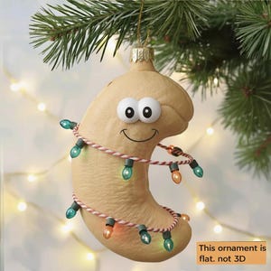 May include: A whimsical cashew-shaped Christmas ornament with a smiling face and googly eyes, wrapped in a string of colorful lights. The ornament hangs from a Christmas tree branch. Text on the image reads: "This ornament is flat. not 3D."