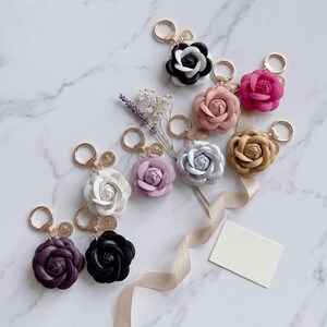 May include: A collection of flower-shaped keychains in various colors, including white, black, pink, purple, and gold. Each keychain features a gold-colored ring and a small charm. The keychains are arranged on a marble surface.