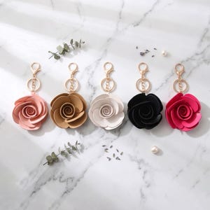 May include: Five rose-shaped keychains in a row, each with a gold-colored clasp and ring. The roses are in shades of pink, tan, white, black, and red. The background is a white marble surface with sprigs of greenery.