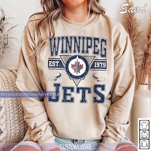 May include: Sand-colored sweatshirt featuring the text "Winnipeg Jets" in navy blue, along with the team logo and the year "1979." The design includes hockey puck graphics. The sweatshirt is a casual style.