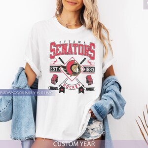 May include: White t-shirt featuring the Ottawa Senators hockey team logo in red, black, and gold. The design includes the team name, crossed hockey sticks, and the text "EST. 1883". The shirt has a crew neck and short sleeves.