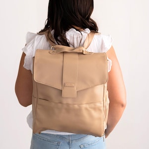 May include: A tan backpack with a flap closure and a top handle. The backpack has a front pocket and a side zipper pocket. The straps are adjustable. The bag is made of a textured material.