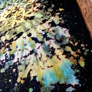 May include: A black t-shirt featuring a bleached tie-dye design with a geometric flower of life pattern. The fabric displays splattered accents in shades of yellow, blue, and soft pink. A small logo with the text Wook Werx Hawaii is visible.