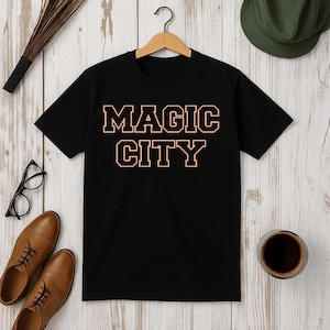 May include: Black t-shirt with the words "MAGIC CITY" in a block letter style with a peach outline. The shirt is hanging on a wooden hanger. The image also includes brown leather shoes, eyeglasses, and a cup of coffee.