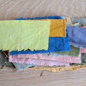 May include: A selection of colourful fabric swatches in shades of yellow, blue, pink, and green. The fabric pieces have frayed edges and are arranged on a light brown wooden surface.