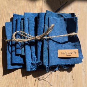 May include: A bundle of indigo blue fabric squares, secured with twine. A small rectangular label with the text "Sunny Side Up" is visible. The fabric has a raw, frayed edge.
