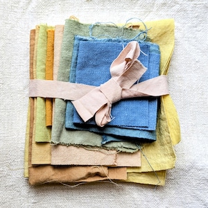 May include: A collection of fabric swatches in a range of colours, including blues, greens, yellows, and browns. The swatches are bound together with a pale pink ribbon. The fabrics display different textures and weights, indicating a variety of materials.