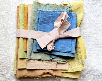 May include: A bundle of fabric swatches in various colors, including shades of blue, green, yellow, and brown. The swatches are tied together with a light pink ribbon. The fabrics have different textures and weights, suggesting a variety of materials.