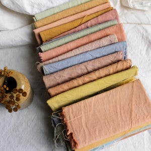 Peut inclure: Un assortiment d'échantillons de tissu aux couleurs pastel, notamment rose, jaune et bleu, disposés sur une surface blanche texturée. Un petit vase en céramique beige avec des fleurs séchées est en arrière-plan.