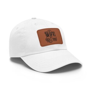 Könnte beinhalten: Weiße Baseballkappe mit einem braunen Lederaufnäher. Der Aufnäher zeigt den Text "MY WIFE WILL KILL YOU" in einer Vintage-Schriftart. Die Kappe hat einen gebogenen Schirm und einen verstellbaren Riemen.